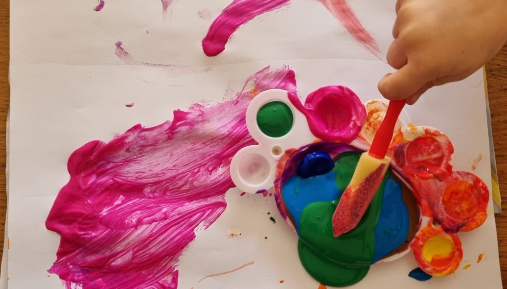 Photo of a piece of paper with bright colours of acrylic paint. A white child's hand holds a foam paintbrush, dipping it into green and blue paint. 